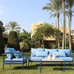 This appears to be an outdoor seating set. It features a blue metal frame with light blue cushions for a sofa and a matching lounge chair, arranged on a grassy lawn with a matching blue coffee table. The scene suggests patio or garden use, with palm trees and a warm, sunny backdrop. Audience: unisex (suitable for both men and women). Notable attributes: bright blue cushions, slim metal frames, modern minimalist style, and a low-profile coffee table with three light-colored vases. Material hints: metal frame and fabric cushions. Color palette: blue cushions and frames, white/cream vases, natural green surroundings.