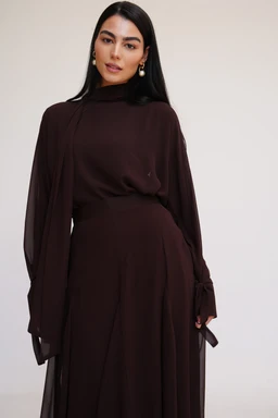 A brown chiffon blouse set on a full-length dress-like top, shown on a female model. The garment features a high neckline, long sheer bishop sleeves with gathered cuffs and tie-back details at the wrists, and a flowing, relaxed bodice that drapes into a matching skirt or layered silhouette. The fabric appears lightweight and slightly sheer with a soft, elegant brown color. Intended for women's fashion, with a modest, formal, and sophisticated style.