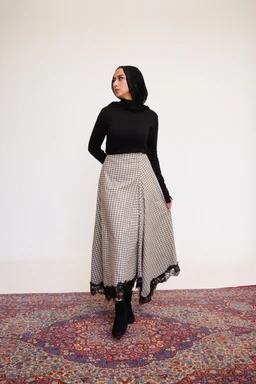 - Description: A plaid lace-trim maxi skirt in beige paired with a black long-sleeve turtleneck top. The skirt has a flowing A-line silhouette with a wrap-style front and a black lace hem.
- Intended audience: Women's.
- Notable attributes: beige and brown plaid pattern, lace edging at the hem, ankle-length flowy skirt, high-waisted fit suggested by the top tucked in, paired with black opaque tights and heeled boots.