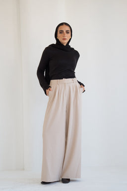 - What it is: Tailored wide-leg trousers in beige, paired with a black long-sleeve turtleneck top and headscarf. The model stands against a plain light background, hands in pockets.
- Audience cues: Women's collection, styled with a modest, contemporary look suitable for a sophisticated, fashion-conscious female audience.
- Notable attributes: Beige/khaki color, high-rise waist with a belt, wide-leg cut, smooth drape, and clean tailored lines. Fabric appears lightweight with a soft, structured finish. Black top provides strong contrast for a classic, minimalist style.