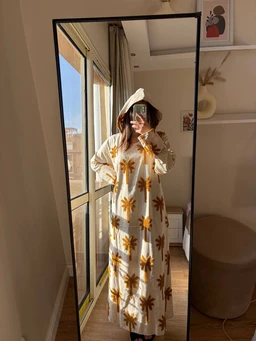 - What the product is: A long, light-colored (likely cream) embroidered dress featuring a palm tree pattern in shades of yellow/gold. The dress has a loose, comfortable fit with long sleeves and a floor-length silhouette.
- Apparent intended audience: Women's (model appears female; styling and fit suggest adult women's clothing).
- Notable attributes: Embroidered palm tree motif all over the dress, light cotton/linen-like fabric, cream base color, long sleeves, ankle-to-floor length, no visible branding, model wearing a headscarf/hood or accessory in a casual indoor setting.