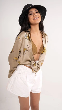 - What it is: A beige button-up shirt featuring tiger embroidery, tied at the waist, worn as a casual top with a bralette underneath. Paired with white high-waisted shorts and a black wide-brim hat.
- Audience and styling cues: Appears designed for women or girls; feminine styling with a relaxed, boho vibe.
- Notable attributes: Light beige fabric with tiger embroidery in darker tones, long sleeves tied at the midsection, cropped length, off-white/white shorts, brown bralette, and a black floppy hat. Material appears lightweight cotton or similar, with a soft, comfortable drape.
