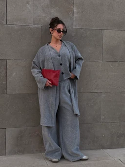 - What the product is: A matching gray loungewear set consisting of a long cardigan or lightweight robe and wide-leg pants, worn together as a coordinated two-piece outfit. The model holds a red clutch.
- Intended audience and styling: Appears to be unisex or women's loungewear, styled for a relaxed, oversized fit with a casual, contemporary look. The model wears dark sunglasses for a chic street-style vibe.
- Notable materials, color, and style attributes: Medium-gray knit/fleece fabric with a soft, drapey texture; long cardigan with a button-down front; wide-leg, relaxed-fit pants; minimal, monochrome styling accented by a red clutch. No visible branding.