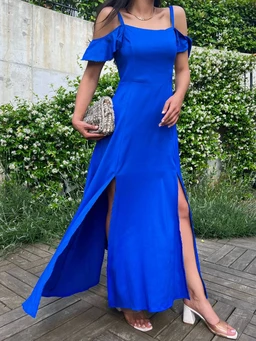 - What the product is: A royal blue, off-the-shoulder maxi dress with short flutter sleeves and a high front slit, designed for a dressy, warm-weather look.
- Apparent intended audience: Women's.
- Notable visible attributes: bright electric blue color, off-the-shoulder neckline with thin straps, fitted bodice that flows into a long skirt with two front slits, lightweight fabric, side-handled small clutch, nude block-heel sandals.