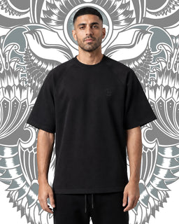 - Product: An oversized black T-shirt with a relaxed fit, short sleeves, and a crew neckline. The shirt appears solid black with a minimal or subtle logo on the left chest.
- Audience and styling cues: Designed for a unisex or men's audience, shown on a male model with a casual, streetwear vibe. The backdrop and styling emphasize a bold, minimalist look suitable for everyday casual wear.
- Materials and attributes visible: Knit fabric with a smooth, matte finish; mid-weight cotton-like material; black color; oversized silhouette as indicated by loose shoulders and extended hem. No branding visible beyond a faint chest mark.