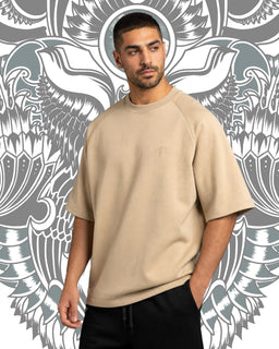 This is an oversized, short-sleeve crewneck sweatshirt in a warm cream/beige color. It appears designed for a unisex or men's audience, featuring a relaxed fit with dropped shoulders and a smooth, fleece-like interior, in a solid, neutral tone. Visible materials suggest a soft knit fabric with a matte finish and minimal detailing, keeping the focus on the garment’s oversized silhouette.