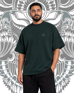 A dark teal oversized short-sleeve T-shirt for men. The model appears adult male, showcasing a loose, relaxed fit with dropped shoulders and a straight hem. Notable attributes include a solid dark teal color, mid-to-long sleeve length, and a small, subtle circular emblem on the chest. The background features a bold, ornamental grayscale pattern. The garment appears to be made of a soft cotton or cotton-blend fabric with a simple, minimalist style.