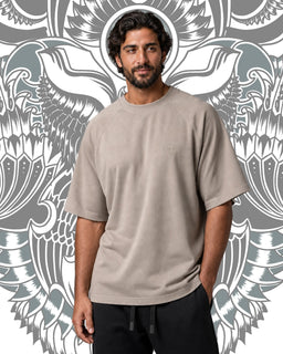 A unisex-looking oversized short-sleeve crewneck sweatshirt in a light taupe/stone color. The model is a man wearing a loose, relaxed fit with mid-length sleeves and a soft, drapey fabric, giving a casual, streetwear vibe. Notable attributes: solid neutral tone, oversized silhouette, and minimal detailing with a subtle drop shoulder.