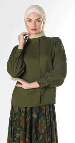 This is a women's blouse, shown on a female model wearing a light-colored hijab. The blouse is an olive green crepe with a high neck, long puff sleeves, and subtle front seam details, pairing a tailored yet relaxed silhouette.
Visible materials, colors, and style: olive crepe fabric, long bishop sleeves with button cuffs, high neckline, clean front with minimal detailing, and a dark floral skirt visible below for styling context.