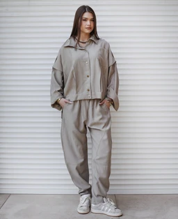 Product is a beige, relaxed-fit linen set consisting of a cropped button-front jacket and matching wide-leg pants. Appears styled for a unisex or fashion-forward casual look, with a loose, oversized silhouette and layered, architectural sleeves. Visible materials suggest light, breathable linen in a soft beige/khaki tone; styling includes a cropped jacket with a hem-aligned waist and elastic or relaxed waistband on the pants, paired with light sneakers.