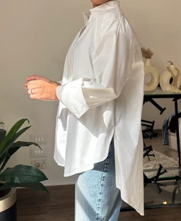 A white oversized shirt with a loose, relaxed fit and drop-shoulder silhouette, styled in a side profile. The model appears to be female, pairing the shirt with light-wash denim jeans for a casual, minimal look; the shirt features long sleeves and a high-low hem with a slightly longer back. Notable visible details include crisp cotton fabric, a subtle stand collar with a small neckline opening, and a clean, minimal aesthetic.