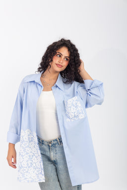 - A light blue, oversized shirt/dress-style top with long sleeves, shown unbuttoned over a white top. The model appears to be a woman, suggesting a women's fit and styling.
- Notable details: white lace/guipure panels on the front pocket and lower left panel, giving a decorative contrast to the solid poplin fabric. The shirt has a classic collar and a freesize, relaxed silhouette.