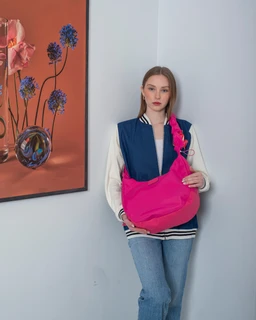 - What the product is: A bright pink everyday bag carried by a woman in a casual casual-teen/young adult look.
- Apparent audience: Women (unisex could be possible, but styling and the model suggest a female audience).
- Notable attributes: Neon pink, soft fabric/nylon-style material, medium-sized hobo/camera-bag shape with a single strap over the shoulder; worn with a two-tone varsity-style jacket and light jeans, bright color contrast against light walls.