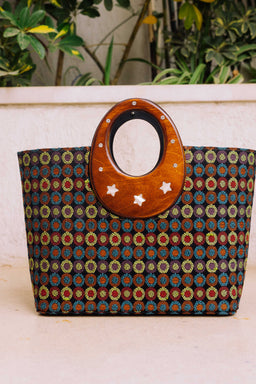 - A patterned tote bag with a circular wooden handle labeled “Black Edition.” The bag features a dark woven fabric with a repeating circular motif in blue, red, and tan tones, set against a matte brown background, and a natural wood ring at the top adorned with small metal studs. The tote is sized for everyday carry and appears suited for women or unisex use, with a lightweight, casual style and decorative star accents on the wooden handle. Notable materials visible include wood for the handle and a textile woven fabric for the bag body, with earthy brown and multicolor patterning.