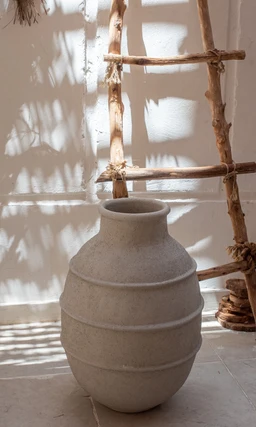 - What it is: A ceramic vase with a rounded, ribbed silhouette and a short neck.
- Intended audience: Unisex (suitable for any interior decor; no gender-specific styling).
- Notable attributes: Matte light gray/stone color, bulbous body with three subtle horizontal rings, narrow neck, and simple, rustic finish. The photo shows the vase placed against a textured wall with natural wood ladder decor in the background.