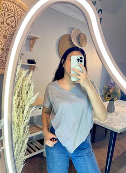 - Product: A casual short-sleeve V-neck T-shirt, styled in a relaxed fit and worn with blue jeans. The shirt hangs loosely with a front tuck to one side, giving a casual, everyday look.
- Audience: Women (based on the neckline, fit, and styling from the model).
- Materials/colors/style visible: Light gray fabric with a soft, smooth texture; solid color; minimalist, contemporary everyday casual style. No branding visible.
