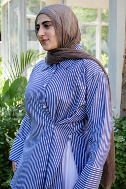 - What the product is: A blue striped button-down shirt, styled with a twist at the waist to create a gathered, wrapped look.
- Target audience: Women's.
- Notable attributes visible: blue and white vertical stripes, long sleeves with button cuffs, collared neckline, front placket with buttons, and a twisted/ruched waist that adds a gathered silhouette. The fabric appears lightweight with a slightly crisp drape. The model is wearing a hijab, suggesting modest styling.