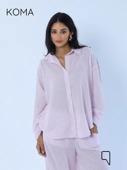 - Product: Matilda Long Sleeves Shirt with striped pattern in soft pink. It features a relaxed, oversized fit with a classic shirt collar and a hidden button front, made from a lightweight, textured striped fabric.
- Audience: Women (as indicated by the model and styling).
- Notable attributes: Light pink color with subtle vertical stripes, long sleeves, oversized/floaty silhouette, collared neckline, and a casual button-down shirt styling. Not showing any branding beyond the visible KOMA label.