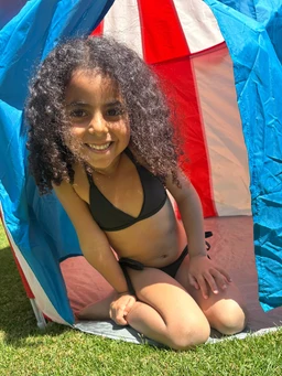 - What it is: A young girl wearing a black bikini set, posing inside a blue pop-up tent on grass.
- Intended audience: Girls (youth) based on the model and sizing.
- Notable attributes visible: Black bikini with simple triangle top and matching bottoms; bright blue tent with red and white panels; outdoor daytime setting with green grass. No brand visible.