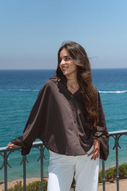 - What the product is: A loose, button-front blouse with a relaxed drape, shown on a female model at a coastal outdoor setting.
- Apparent audience and styling: Designed for women; oversized, billowy silhouette with wide sleeves and a casual, breezy vibe suitable for warm weather.
- Notable attributes visible: Burnt brown color, lightweight, flowy fabric with a soft sheen, V-neckline, button placket, and a slightly cropped/hip-length hem paired with white bottoms against a beach backdrop.
