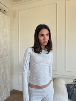 This is a long-sleeve, form-fitting top and matching bottom set in heather light gray. The model appears to be a woman; the styling is minimalist and fitted, with a crew neckline and ruched sides creating a slim silhouette. The fabric looks soft and stretchable, likely a jersey or knit blend, in a solid light gray color. The overall look is casual loungewear or everyday wear with a clean, monochrome style.