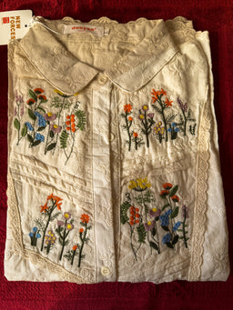 - What the product is: A vintage ivory/cream embroidered blouse with floral embroidery panels on the front.
- Apparent audience: Women (adult sizing inferred from the blouse cut and styling, including a collared neckline and button-down front).
- Notable attributes: Lightweight cotton or cotton-blend fabric with colorful floral embroidery in orange, yellow, red, blue, green, and purple on the front panels; collared neckline, button closure, scalloped/eyelet trim along seams, and decorative stitching. The overall look is delicate, vintage-inspired with a pleated or panel-stitched front.