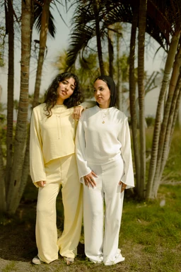 Two models wearing matching loungewear sets in solid pastel colors. The left outfit is a pale yellow two-piece set with a long-sleeve top and loose, wide-leg pants; the right outfit is a white two-piece set with a long-sleeve top featuring a subtle neckline and matching wide-leg pants. Both looks have a relaxed, cozy style suitable for women, with draped silhouettes and soft, breathable fabrics. The overall styling suggests comfortable, casual homewear or lounge attire in a tropical outdoor setting.