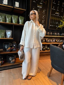 - Product: Crushed linen set in creamy color, shown as a matching loose-fit top and wide-leg pants.
- Audience: Women, based on feminine silhouette, styling with a hijab and accessories.
- Notable attributes: Creamy crushed linen texture, oversized relaxed fit, long-sleeve top with a collar, wide-leg cropped pants, minimalistic sandals, and a small handbag. The look is casual, comfortable, and polished with a neutral monochrome palette.