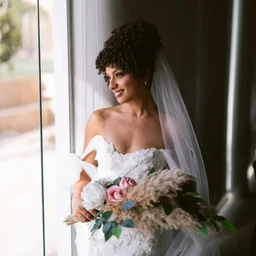- Product: Bridal package item featuring a bridal look displayed on a female model.
- Audience: Women (bride) based on the off-shoulder wedding gown, veil, and bouquet styling.
- Visible attributes: White lace off-shoulder wedding dress with a long veil; curly short hairstyle; bouquet with white and pink roses, greenery, and pampas-like beige accents; soft natural light by a window in an indoor setting. No branding visible.