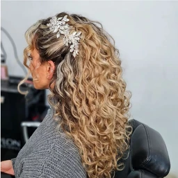 This appears to be a formal or party-inspired hairstyle rather than a clothing item. The model has long, voluminous, tight curls with a light brown/blonde balayage and a sparkling decorative hairpiece featuring leaf-like crystal embellishments. The look is styled for a feminine audience, with a romantic, soirée-ready vibe and emphasis on shimmering accessories.