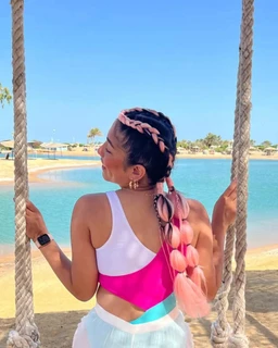 - What it is: A bikini/swimwear top in a color-block design worn by a woman, shown from the back and side while standing on a hanging rope swing near a beach.
- Intended audience: Women's or unisex styling intended for female-presenting individuals.
- Notable attributes: Bright color-block palette with white, pink, and light blue/teal accents; braided pink hair with a long fishtail and a decorative pink braid; casual beach setting with a sleeveless high-cut back; medium to long hair styled in multiple braids; overall sporty-yet-stylish beachwear vibe.