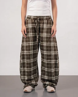 - Product: Plaid wide-leg trousers in brown tones.
- Audience: Unisex or ambiguous gender presentation (loose, relaxed fit with a casual styling; model appears neutral).
- Visible features: Brown and beige plaid print, elastic waistband with drawstring, very loose, wide-leg silhouette that drapes to the floor, cuffed at the ankles, paired with a white top and casual sneakers. Notable materials and styling: soft, drapey fabric with a casual, lounge or pajama-trouser vibe; earthy brown plaid pattern.