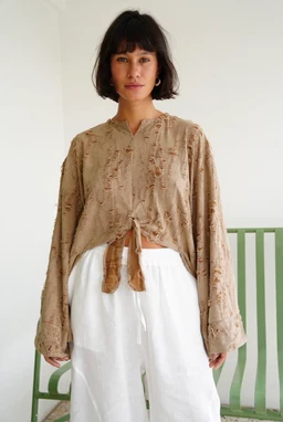 - A lightweight, long-sleeve blouse with a fringe texture and a natural beige/tan color. The top features a V-neck with a tie-front knot and a relaxed, slightly oversized fit, paired with white pants in the photo.
- Intended audience: women's.
- Notable attributes: soft, textured fabric with fringe detailing, casual earthy tone, loose silhouette, and a cropped front tie that creates a relaxed, boho-inspired look.