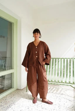This is a brown Balloon Pants set featuring a matching top and wide, voluminous leg pants. The styling appears unisex or leaning toward a relaxed women’s fit, with a loose, draped silhouette and a tie-front detail on the top. Notable attributes include earthy brown fabric, drop-shoulder sleeves, and a lightweight, breathable material with a matte finish.