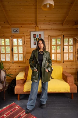 - What the product is: A knee-length, olive-green leather jacket styled for casual outerwear, shown worn by a female model.
- Apparent audience and styling: Targeted to women; the model wears it over a dark top with wide-leg jeans and black shoes, styled with layered necklaces for a relaxed, urban look.
- Notable visible attributes: Smooth, glossy leather with a slightly oversized fit, large lapels, and zippered pockets; dark interior lining peeks at the sleeves, in a warm, wooden room setting with yellow seating in the background.