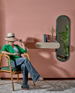 - What the product is: A fashion-oriented photo featuring a woman seated in a modern setting; the item highlighted appears to be a lightweight, stylish outfit accessory and environment rather than a single clearly defined product.
- Apparent audience: unisex-leaning women’s styling, based on the model and styling cues (hat, green cardigan, wide-legged jeans, open-toe sandals).
- Notable attributes visible: the model wears a light straw hat with a black band, a bright green cardigan over a white top, and blue wide-leg jeans. The setting features a soft pink wall, a wooden chair, and a curved wall-mounted shelf with books and a small plant reflected in an oval mirror-like panel; the overall color palette is pink, green, and natural wood tones. Style impression is casual-meets-chic with relaxed, summery vibes. The material textures visible include fabric clothing and a smooth, matte wall finish.