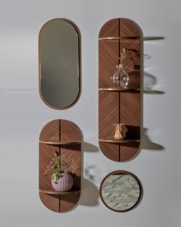The photo shows a modular wall shelving set with a mid-century feel. It includes two tall oval-edged brown wood shelves with vertical split accents, a smaller matching oval shelf, and a round mirror with a wood-toned frame. The pieces are mounted on a light gray wall, with decorative items (vase with flowers, glass vase, and a small pot) arranged on the shelves. The overall style is warm wood tones and rounded shapes, giving a unisex appeal. Notable materials and colors: warm brown wood veneer with a peach-brown frame; glass mirror; muted gray-green reflective surface in the round mirror.