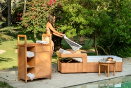 This is an outdoor bathroom set shown by a woman in a resort-like garden setting. It appears intended for adult women or unisex use, featuring a wooden bath vanity with shelves and a towel stack, a matching bathtub with a raised side shelf, and a small side table. Notable materials include light-to-medium stained wood with fabric towels in white and gray, and a reclining seating or towel-drying area integrated into the tub unit, all in a cohesive natural wood style.