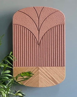- What the product is: A pink-toned, decorative wall hanging or plate-like piece with a geometric carved pattern, likely ceramic or wood composite.
- Apparent audience: Unisex (suitable for any gender), based on neutral pastel color and contemporary design.
- Notable attributes: Two-tone design with a blush pink upper section featuring a stylized, radiating leaf/vein motif, and a lower beige-wood tone with diagonal, chevron-like grain. Textured surface with vertical grooves in the pink portion and smooth, curved edges, mounted on a gray wall.