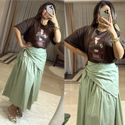 - A casual women’s outfit featuring a dark brown graphic T-shirt paired with a light sage-green wrap-style midi skirt. The model is wearing layered bracelets and neutral heels, with a relaxed, oversized tee and a draped, tied-front skirt for a breezy, urban look. Notable attributes: dark brown short-sleeve T-shirt with a printed graphic, light sage green wrap/skirt with asymmetric draping and ruching at the waist. Material and color cues visible: soft cotton tee, lightweight woven skirt in a muted green tone, and simple metallic footwear accessories.