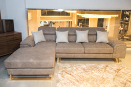 - What the product is: A modular L-shaped sofa with a matching chaise, upholstered in light brown/stone fabric.
- Apparent audience: Unisex (suitable for men or women), shown as a large sectional for a living room.
- Notable attributes: Light brown/taupe fabric, quilted seam details on the arms, five light gray throw pillows, gold-toned metal legs, chaise on the left, contemporary low-profile design.