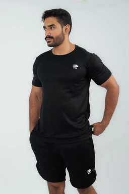 A plain black short-sleeve t-shirt styled on a male model. Intended audience appears to be men, judged by the fit and cut; the shirt is solid black with a small light-colored logo on the left chest. Notable details include a crew neck, fitted yet comfortable silhouette, and a matching pair of black shorts shown for a coordinated athletic/casual look. Material appears to be a smooth knit, likely cotton or cotton-blend, with a matte finish.