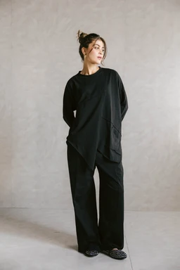 - A black unfinished cotton set displayed on a female model. The outfit consists of a loose, oversized long-sleeve top with an asymmetric hem and matching wide-leg pants. 
- The model appears to be adult female; styling is minimal and relaxed with hair in a casual bun, suggesting a unisex or androgynous aesthetic. 
- Visible materials and colors: solid black cotton, matte finish, with a soft drape. Notable features include the asymmetrical panel on the top and roomy, cropped-length sleeves, plus wide-leg trouser silhouette. No branding visible.