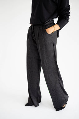 A pair of black satin tailored pants with a wide-leg silhouette. The model appears to be female, wearing a black top; the pants have a high rise with a relaxed, fluid drape and soft sheen, suggesting a satin fabric. Notable details include a subtle texture or pattern in the fabric, clean front with side pockets, and a cropped heel visible for styling. Material emphasis: black satin/sateen with a tailored, pant-ereal cut. Color: solid black. Style: wide-leg, high-rise, tailored pants. Audience: women.