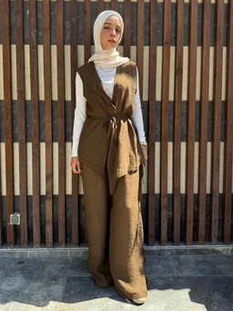 This is a full-length fashion photo featuring a woman wearing a sleeveless, wrap-style brown linen vest over a white long-sleeve top and a matching brown wide-leg pant set, cinched at the waist with a tie. The look appears to target adult women, styled modestly with a light hijab in a neutral tone, suggesting a contemporary, airy linen fabric with earthy brown color. Visible attributes include the textured linen material, waist-tie belt, soft drape, and neutral-toned hijab against a striped wooden backdrop.