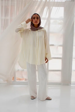 The image shows a loose, flowing striped poplin blouse in a soft, light ivory or cream color, paired with matching wide-leg pants. It appears aimed at a women’s audience, with modest, relaxed-cut styling and a lightweight, airy look. Notable details include the relaxed silhouette, long sleeves, subtle vertical stripes, and a soft, luminous fabric that reads as breathable poplin.