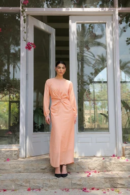 - What it is: A long, peach-toned knit/soft-fabric dress with a twist knot at the waist, full-length, with long sleeves.
- Intended audience: Likely women's fashion.
- Notable attributes: Peach color, long sleeves, ankle-length, gentle drape, twist/knot detail at the midsection, simple crew neck, paired with black flats. Material appears soft and comfortable, with a smooth, matte finish. The styling is clean and modest, photographed on a model standing on steps in front of a light-colored door and glass windows.