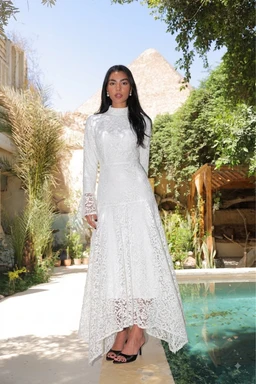 - What the product is: A white lace dress with a high neckline and long sleeves, featuring an intricate floral lace pattern and a floor-grazing to ankle-length asymmetrical hem.
- Apparent audience: Women (adult female model, feminine styling and delicate lace silhouette).
- Notable attributes: White lace fabric, high neck, long sleeves, fitted bodice with a flowing skirt, scalloped/pointed hem with an asymmetrical length, delicate floral pattern, sheer long sleeves, and a subtle layering/lining for coverage.