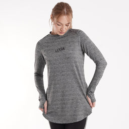 - What it is: A heather gray, long-sleeve crewneck top with a relaxed tunic-length hem. The shirt has a slim, minimal silhouette and a small text logo centered on the chest.
- Intended audience: Women's (judging by the fit, length, and styling).
- Notable attributes: Material appears to be a soft, knit jersey in gray marle; long sleeves with fitted cuffs; slightly curved, tunic-style hem; simple, black text logo on the chest. No visible branding besides the small chest print.
