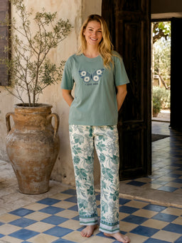 - Product: Short-sleeve pajama set (top and long pants). The top is a light teal T-shirt with a playful graphic on the chest; the pants are white with a teal-blue botanical/leaf print and a matching teal border at the cuffs.

- Audience and styling: Intended for women; the relaxed fit, soft fabric look, and styling with bare feet and a casual, home lounging vibe suggest loungewear for a feminine, comfortable at-home wear.

- Materials and colors: The visible colors are light teal for the top and white with teal botanical print for the pants; the overall look is soft, breathable-feeling cotton or cotton-blend fabric with a casual, cozy pajama style.